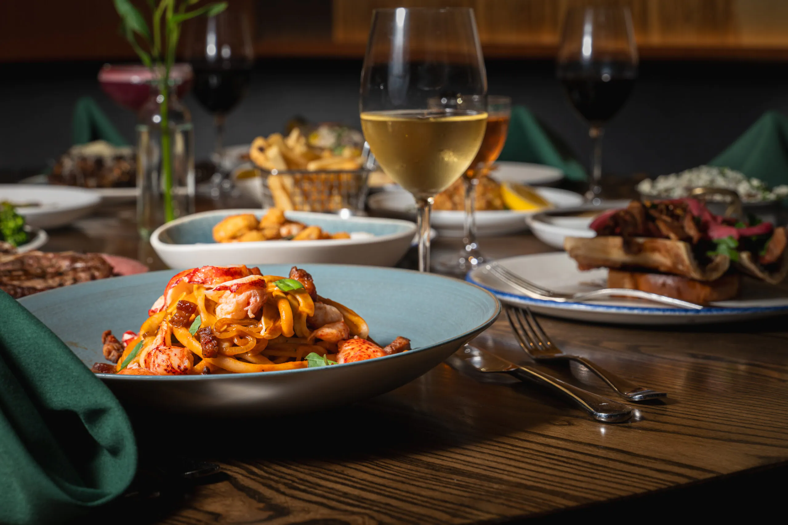A plate of pasta with seafood sits on a wooden table, surrounded by wine glasses and other dishes in a restaurant setting.