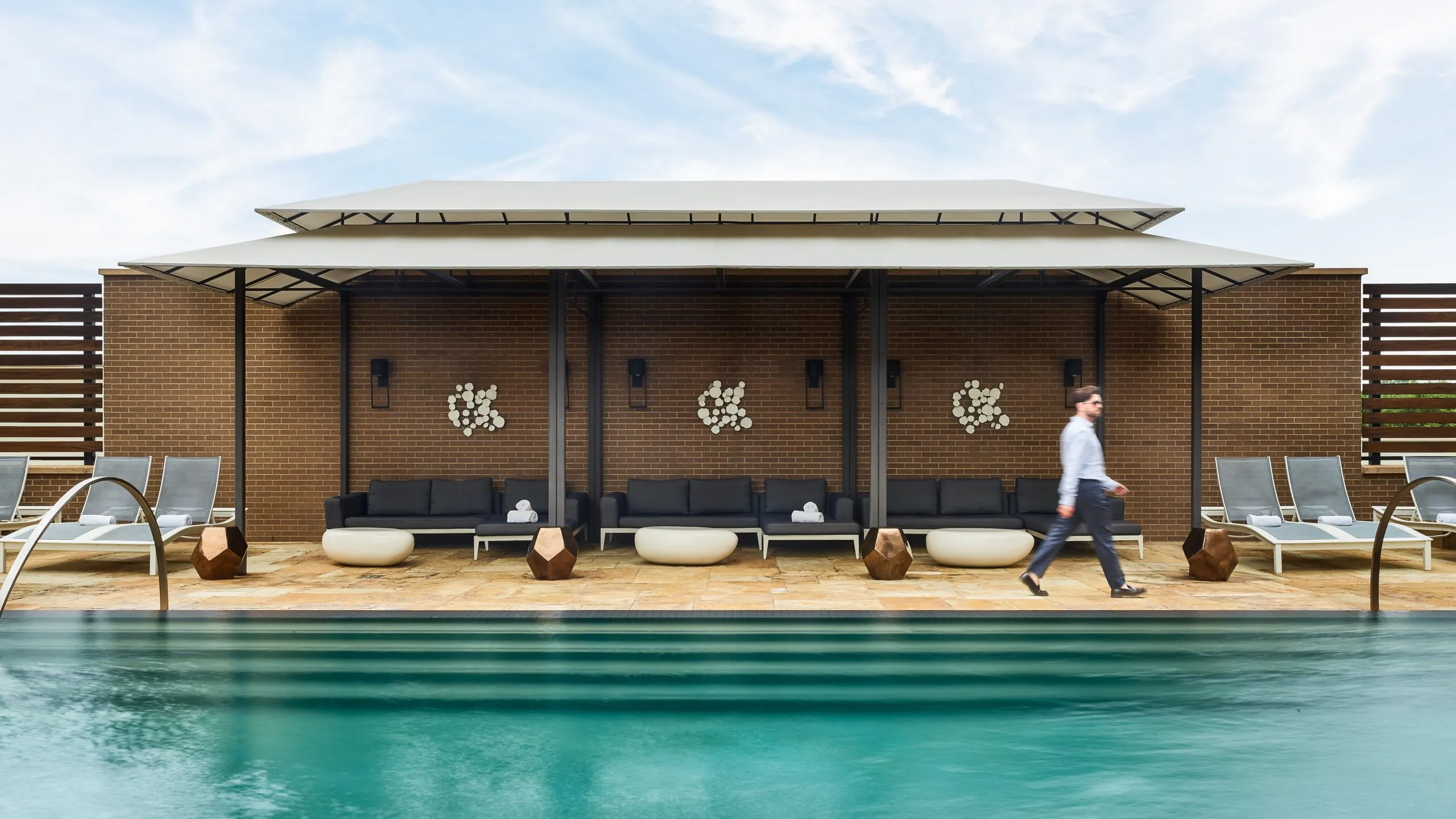 A man walks by a modern pool with lounge chairs and shaded seating area against a brick wall under a partly cloudy sky.