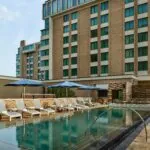 Outdoor pool with lounge chairs and blue umbrellas beside a modern hotel building, with large windows and stone patio.