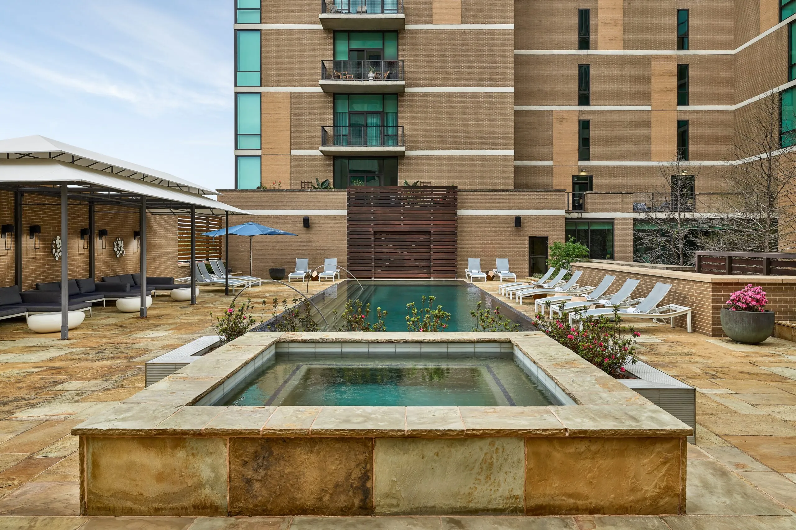 Modern outdoor pool area with lounge chairs, shaded seating, a hot tub in front, and a tall apartment building in the background.