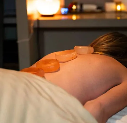 Person lying face down on a massage table with hot stones placed on their back in a dimly lit, relaxing spa setting.