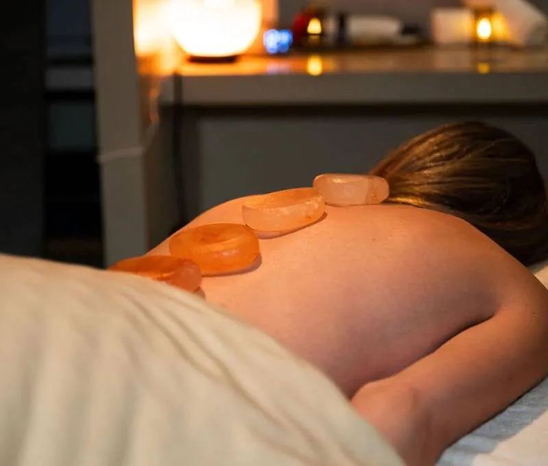 Person lying face down on a massage table with hot stones placed on their back in a dimly lit, relaxing spa setting.