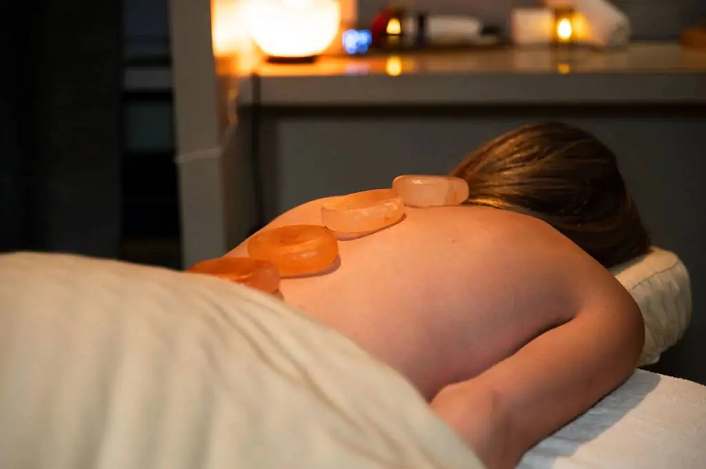 Person lying face down on a massage table with hot stones placed on their back in a dimly lit, relaxing spa setting.