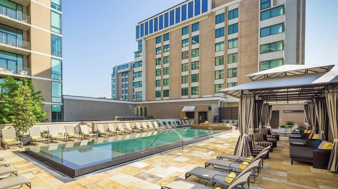Outdoor pool with lounge chairs, cabana seating, and tall modern buildings in the background under a clear blue sky.