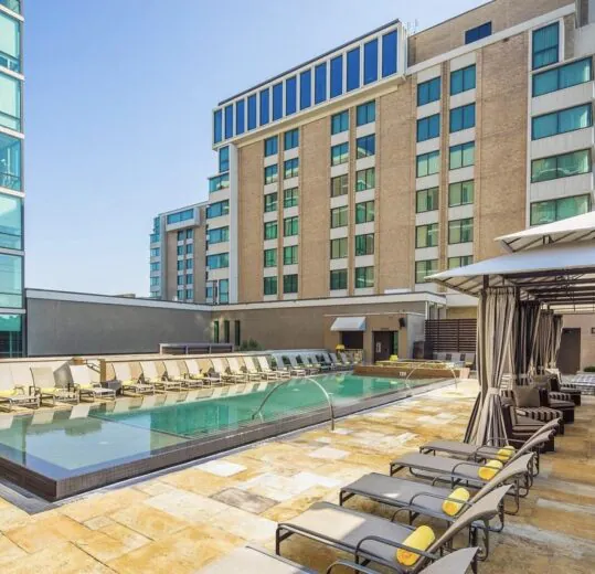 Outdoor pool with lounge chairs, cabana seating, and tall modern buildings in the background under a clear blue sky.