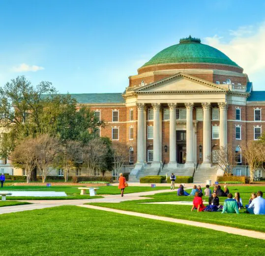 People sit and walk on a grassy lawn in front of a large brick building with columns and a green domed roof under a clear blue sky.