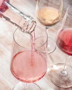 Close-up of rosé wine being poured into a glass, surrounded by other filled wine glasses on a light wood surface.