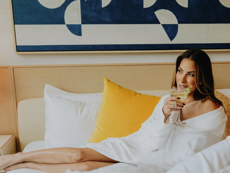 A woman wearing a white robe lounges on a neatly made hotel bed with white linens and a bright yellow pillow. She holds a martini glass and smiles thoughtfully, with modern blue-and-white abstract artwork hanging on the wall behind her.