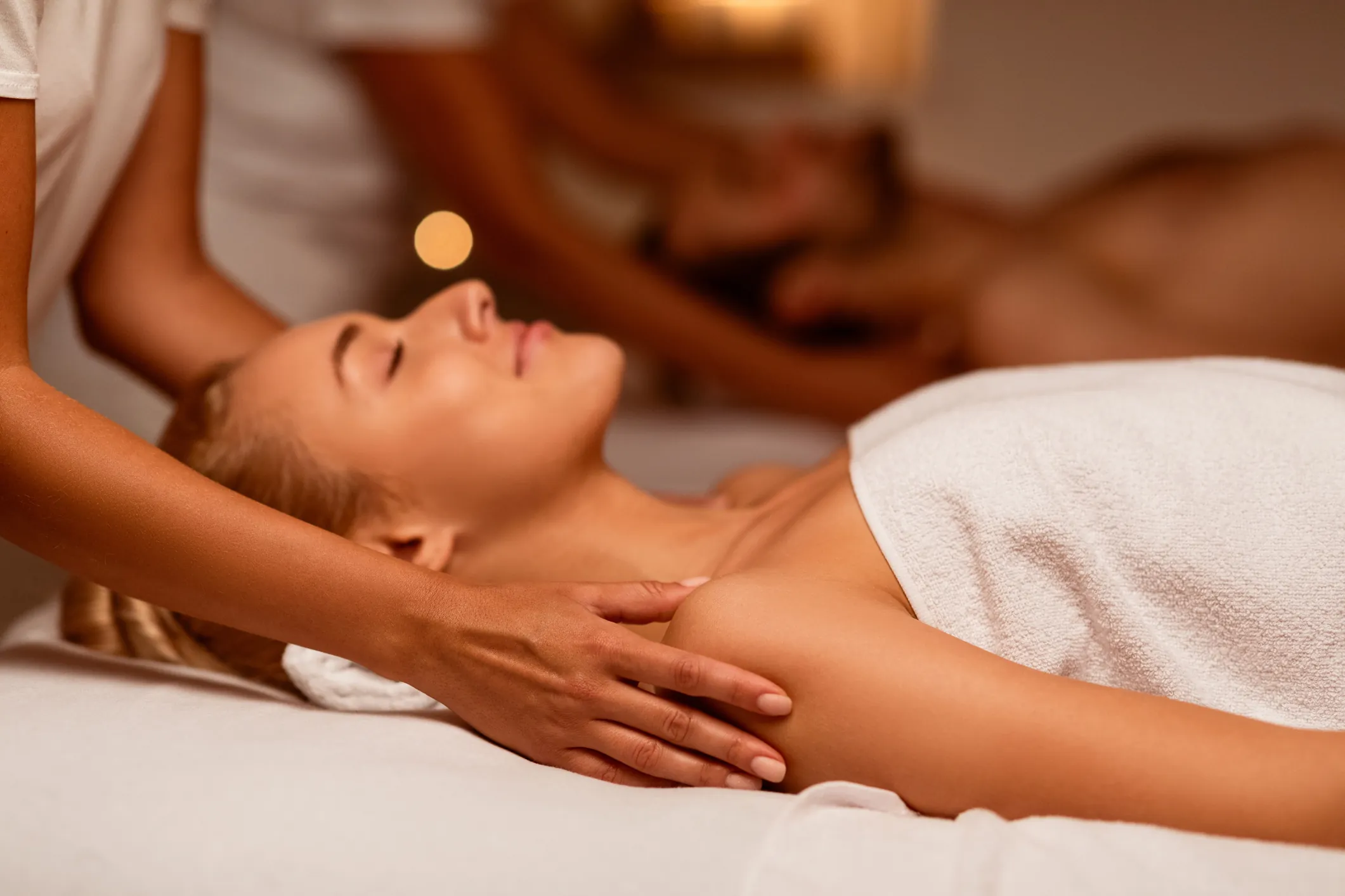 Woman receiving a shoulder massage at a spa, lying on a massage table with a towel, another person receiving a massage in the background.