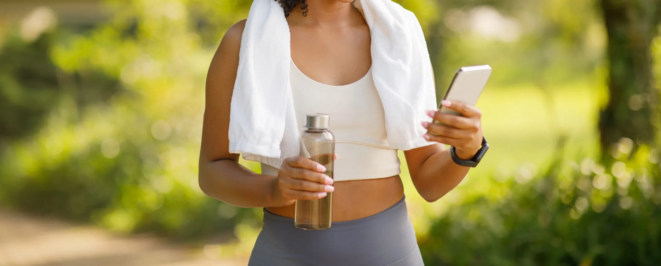 Woman in workout clothes with a towel around her neck holds a water bottle and checks her phone outdoors in a green, sunny environment.