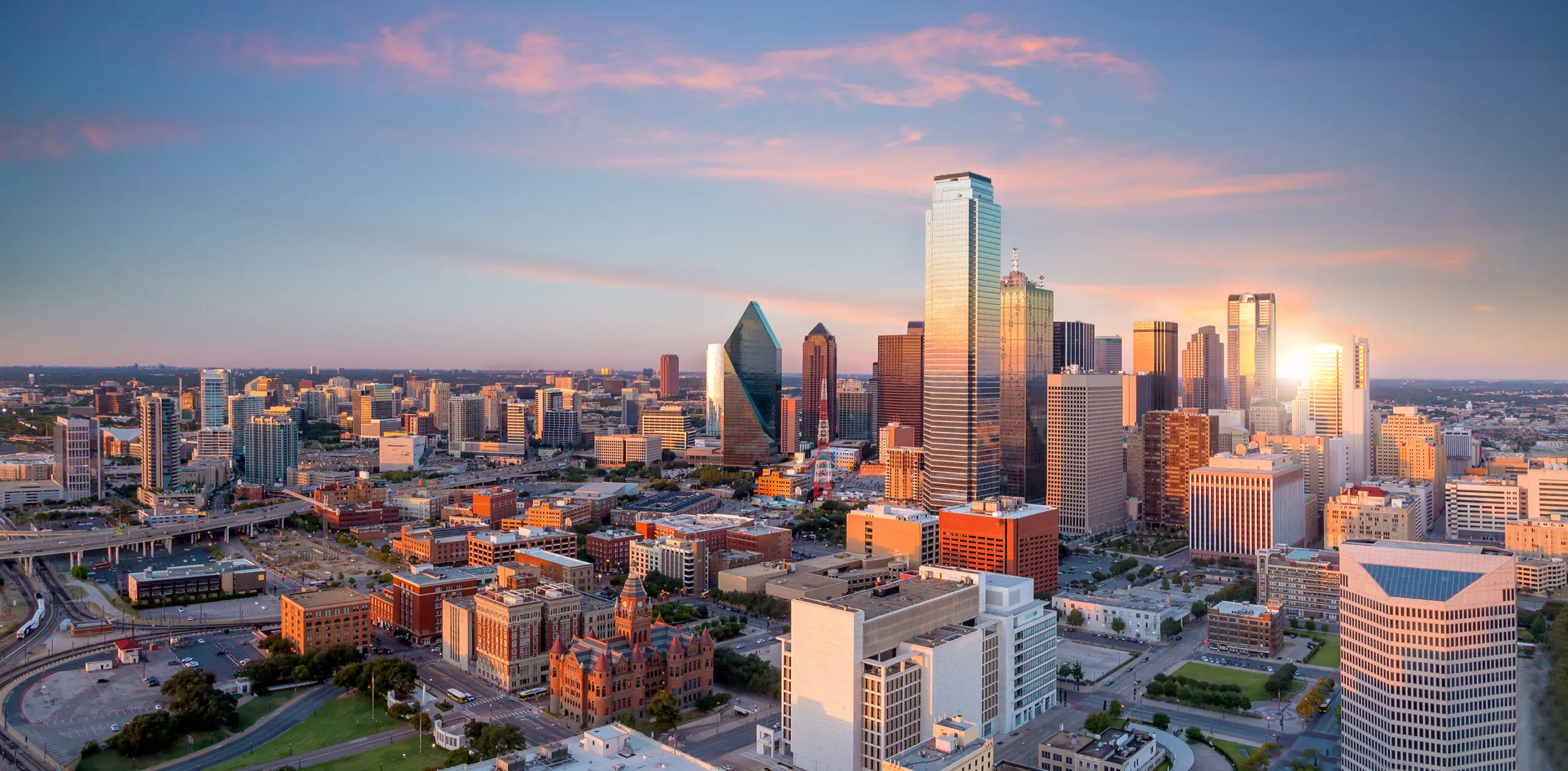 Downtown Dallas skyline at sunset with modern skyscrapers, historic buildings, and a pink sky in the background.