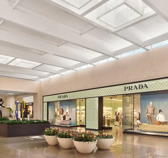 Upscale shopping mall interior with a large Prada storefront, mannequins in display windows, potted plants, and statues visible in the background.