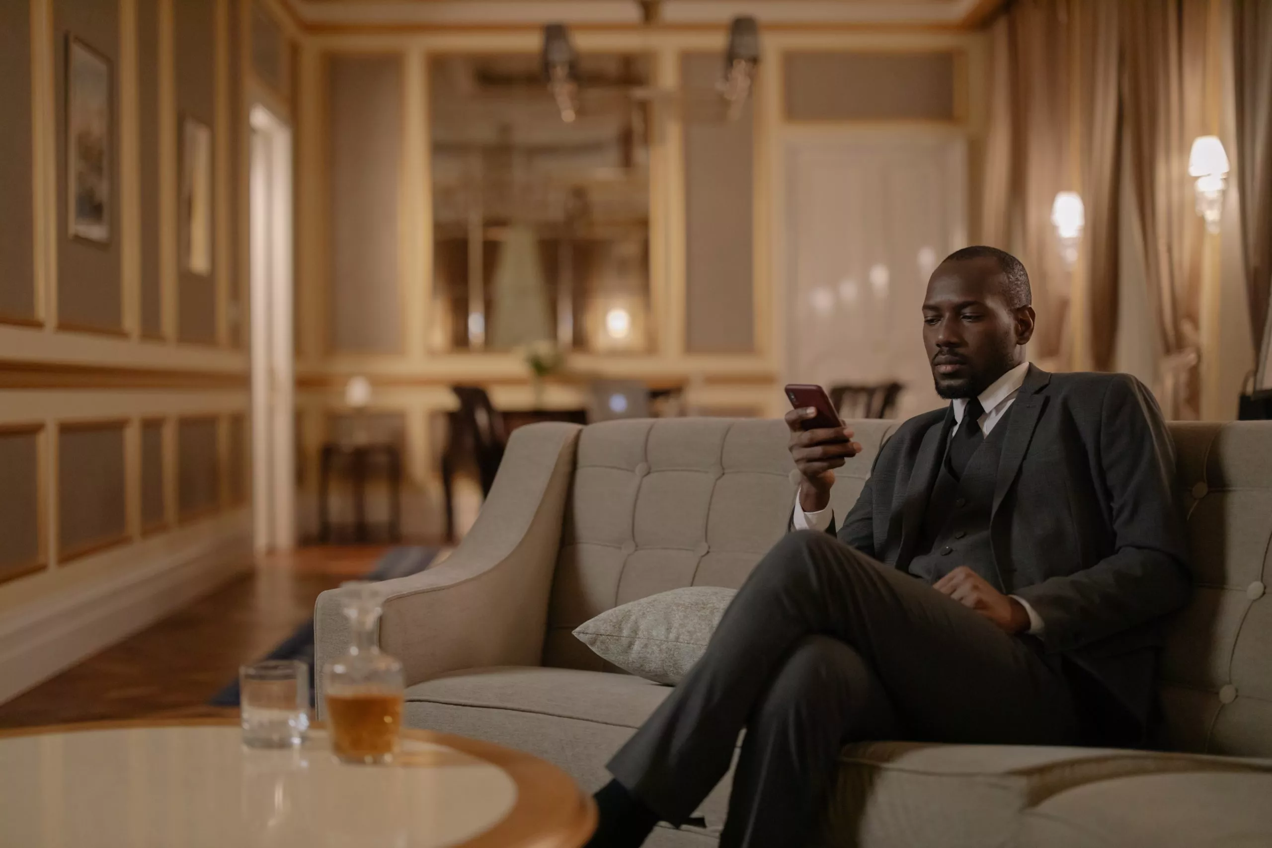 Man in a suit and tie relaxing on a gray lounge couch on his cell phone with a cocktail in the Highland Dallas hotel lobby
