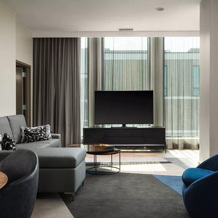 Living room in a King Suite with a gray sectional couch with a blue chair and wooden coffee table and a gray entertainment center with a large flat screen tv and floor to ceiling windows with gray drapes in Dallas, TX